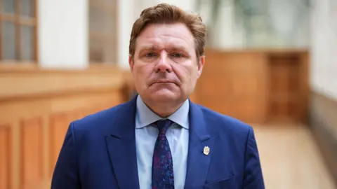 PA Media Birmingham City Council leader John Cotton stands in a corridor lined with wooden panels. He is wearing a navy blue suit, blue shirt and purple and blue tie and has a serious expression. He has light brown hair combed back from his face.