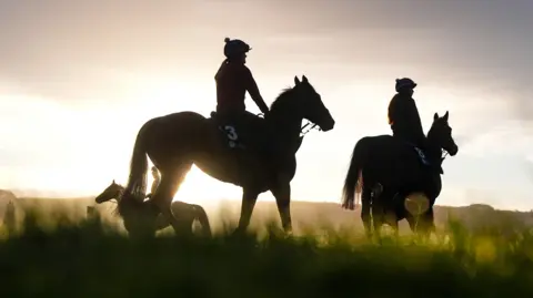 A silhouette of three horses and their jockeys standing in a field of grass at sunset. The light is soft and is coming from directly behind the horse.