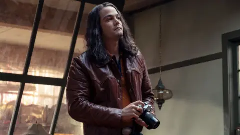 Netflix Daniel Zovatto como Rodney Alcala em Mulher da Hora. Ele tem cabelo comprido, uma jaqueta de couro cor de vinho e segura uma câmera.