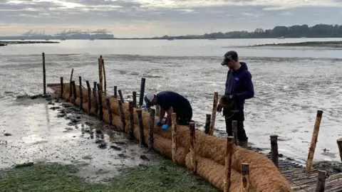 Two men work on barrier along a walkway that heads out into a saltmarsh. The Port of Felixstowe can be seen in the distance on a cloudy day. 