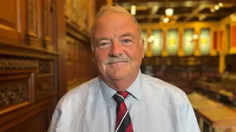 Martyn Perkins is wearing is wearing a light blue shirt, and a red, white and black tie. He's standing in the House of Keys chamber.