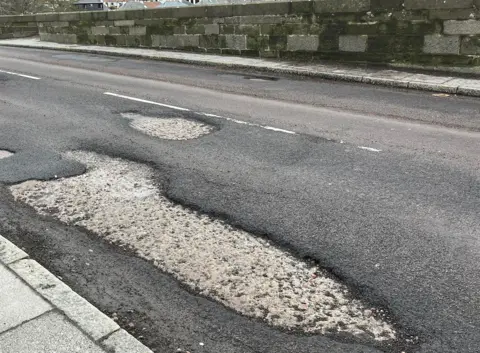 A tarmac road across a bridge with large areas of where the surface has been damaged