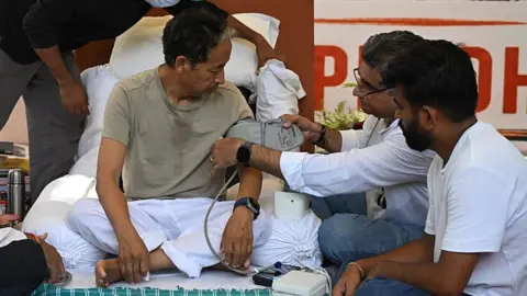 Cockroach Janta Party (CJP) founder Abhijeet Dipke (R) watches as doctors check activist Sonam Wangchuk's vital signs during his hunger strike in New Delhi on July 15, 2026, demanding the resignation of India's Education Minister Dharmendra Pradhan over alleged examination irregularities.