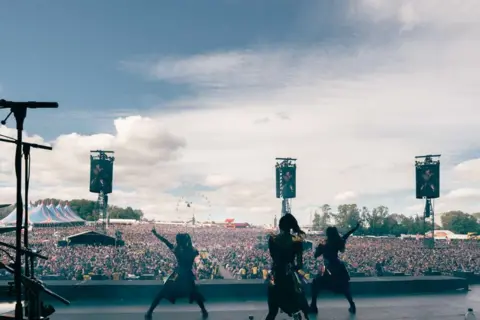 Roger Alarcon/Download Festival Three women on a stage from behind, looking out to a large crowd while dancing. 