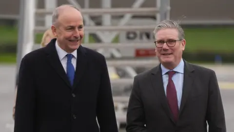 Brian Lawless/PA Micheal Martin, a man with grey hair, a black coat, and blue tie and Sir Keir Starmer, a man with grey hair, glasses, grey suit and claret tie at the airport in Cork.
