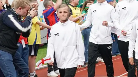 Marcus Tate/ Strike Global Limited A teenage boy wearing a white sports kit walks along an athletic track with other football players. On the sidelines, a crowd cheers the team on. He has blonde hair, blue eyes and a birthmark across one side of his face and neck.