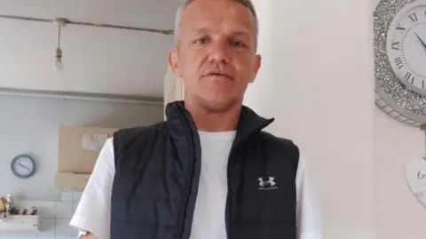 Peter Phillips has close cropped grey hair and is wearing a black sleeveless jacket with a white t-shirt underneath. He is standing in a domestic kitchen.