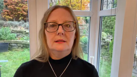 Pauline Jones - A middle-aged fair-haired lady, wearing a achromatic  top, necklace and glasses.