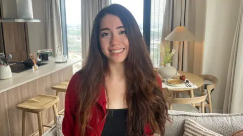 Anna Navabi stands in the living space of a One Eastside apartment, smiling at the camera. She has long dark hair and is wearing a red cardigan over a black top. Behind her is a modern open-plan kitchen and dining area with large windows showing a view over Birmingham.