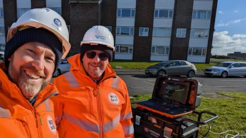 Two Southern Water team members smile for the camera next to a block of flats. They wear orange jackets and white hard hats. A high-tech laptop sits on top of equipment behind them.