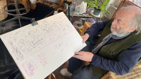 Quentin Blake Sir Quentin holds a white canvas showing a basic sketch of the mural. He has silver hair and beard and wears a blue and green outfit. 