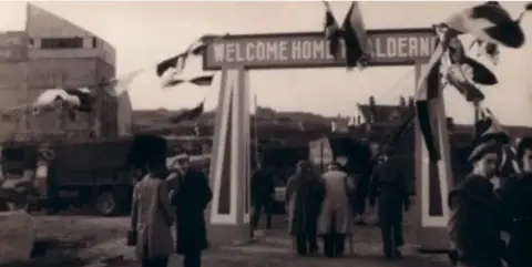 A picture of a large banner saying 'Welcome Home Alderney'. There is bunting, other flags and people walking beneath the banner.