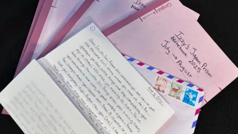 A pile of pink A4 notebooks, the top one titled: Izzy's Japan Prison Notebook 2025 July to August. On top is an envelope postmarked from Osaka, Japan and a letter starting, Dear Iz, oh Iz! Read this when you feel low