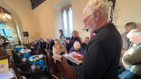 A man holds a small book and looks down at it. Three bells with ropes lie in front of him. In pews nearby, people in winter jackets sit and watch the proceedings. People are stood at the side, as the church is so busy.