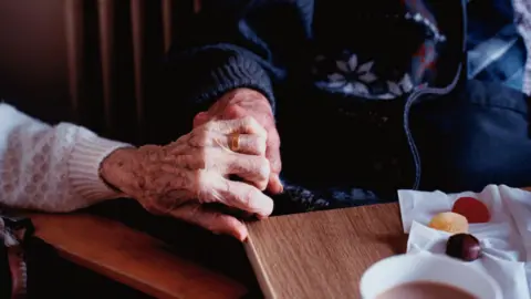 Elderly couple holding hands. They are sat in chairs at a table. There is a cup  of tea on the table and a radiator behind them.