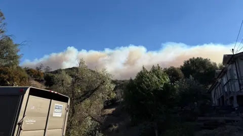 Contributed Thick smoke billows above a large hillside against a blue sky. Trees line the hillside. A horse trailer can be seen to the left of the image.