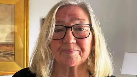 Jo Abbott Jo Abbott with short blonde hair, black-rimmed glasses with the side of a picture and lamp in the background