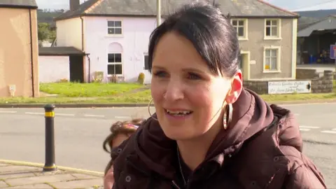 Woman in street talking to journalist. She has dark hair tied back and wears large hoop earrings and a brown coat