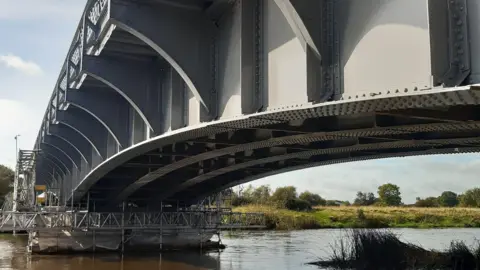 A large steel bridge with an arched shape, going over a river with grassland in the background.