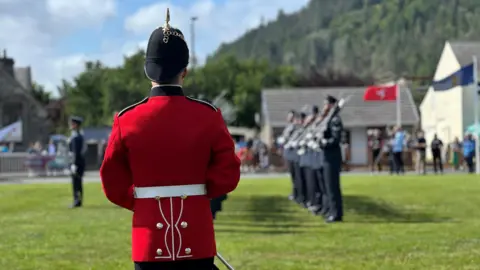 The back of a trumpeter dressed in red, who is facing the Guard of Honour, which is lined up on the green in front of him.