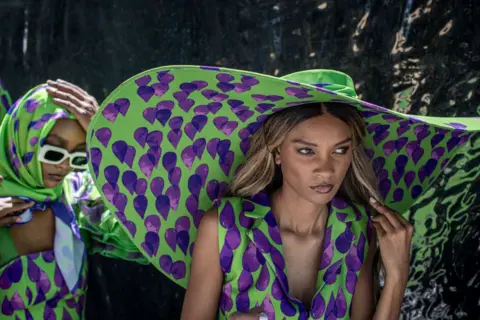 PERS-ANDERS PETTERSSON / GETTY IMAGES Models wait backstage wearing huge hats and clothing featuring the same green-and-purple print.