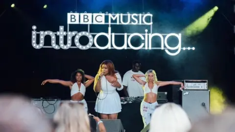 Sisi on stage for BBC Introducing. She is wearing a short white dress and has long dark hair. She has two backing singers, one on either side, wearing white halterneck crop tops and trousers with a man working music decks behind. Over their heads is an illuminated sign reading BBC Music Introducing in a variety of funky fonts.