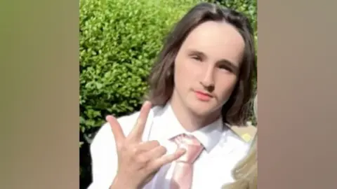 Spencer Crofts is looking at the camera, making a gesture to the camera with his right hand. He is wearing a white shirt and salmon pink tie and has shoulder length brown hair.