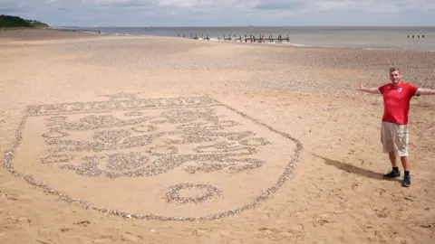 A large England crest made out of stones on a beach. Next to it is a man wearing shorts and a red England top. He has is arms out and is smiling. The sea is behind him.