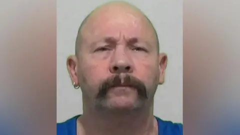 Mugshot of Anthony Cocking. He is bald with a thick dark handlebar moustache, a gold ring in his right ear and is wearing a bright blue shirt