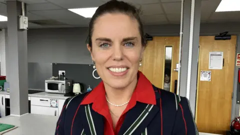 John Rankin headteacher Flora Cooper is standing in front of a kitchen island with a microwave and small chest fridge behind it. She's wearing a dark blue blazer which has red, white and green vertical stripes on it. Smiling broadly she has a red open neck shirt underneath the jacket.