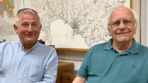 Richard sits on the left in a light blue shirt. Reg is on his right, he is wearing a green polo shirt and has glasses. They both have white hair.