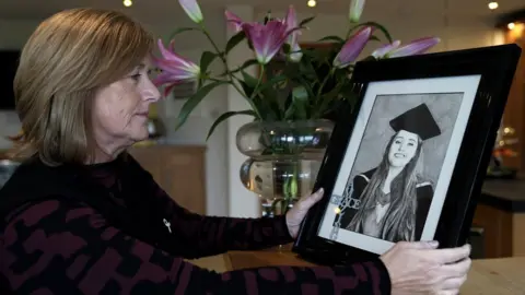 John Fairhall/BBC Gillian Millane has short brown hair. She is wearing black and holding a framed picture of Grace at her university graduation.
