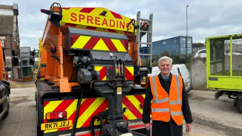 Councillor Trevor Muten (a man with grey hair and a black suit with an orange hi-vis jacket) with one of the new gritting lorries