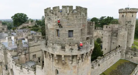 Building restoration specialists abseil down the walls of Warwick Castle's south front, known as Caesar's Tower, as they removed weeds and tidied the 800-year-old stonework during the landmark's annual external clean Two people dressed in red tops and black trousers abseil down the tower of a medieval castle. The picture has been taken from above