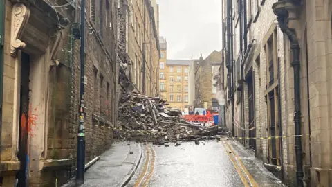 BBC Rubble and roof timbers in a pile blocking a road with double yellow no-parking lines leading up to the scene 