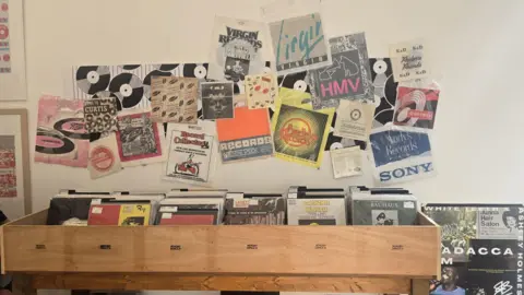 Hayley Coyle/BBC A wooden record bin filled with vinyl albums sits against a wall covered in a collage of vintage record sleeves, labels, and flyers. The wall display includes designs featuring logos such as “Virgin Records,” “HMV,” and “Sony,” alongside black-and-white record graphics and colourful retro artwork. Rows of vinyl records with visible covers are arranged in the bin below, creating a layered, music-themed display.