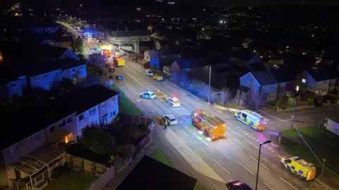 A nighttime aerial view shows a residential street filled with emergency vehicles with flashing blue lights. Several police cars, ambulances, and a fire engine are positioned along the road, which is cordoned off. Houses line both sides of the street, and the wider neighbourhood is visible in the background.