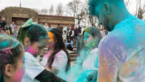 Caroline James Photography A group of people wearing white t-shirts throw bright coloured powder on each other as part of Holi celebrations in a park. They are all smiling.