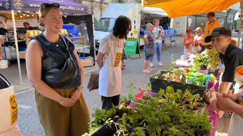 People stand around a market stall displaying trays of herbs, salad leaves and vegetable plants under blue and yellow canopies. Potted plants are arranged on tables covered with purple cloths, while several children wearing aprons help at the stall. Other market traders, shoppers and stalls can be seen in the background, including a coffee stall and a sign for Shambles Market.
