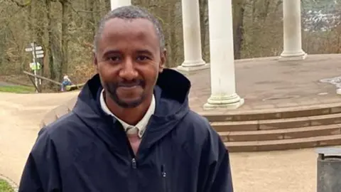 A head and shoulders image of Yonas Kedir. He is wearing a blue waterproof jacket - a white collar can be seen peaking though the top. He is in a park and some columns of a bandstand can be seen behind him.