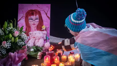 PA Media A child leaves a candle at a tribute for Brianna. Flowers are on the ground, with a pink teddy and pink drawing of Brianna