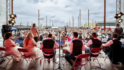 TradFest A photo taken from behind a band on stage. The band is sitting and holding instruments including the fiddle and drums. A huge crowd is dancing. Some have their arms in the air. It's cloudy. 