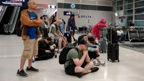 Getty Images A group of people sit on the floor in an airport while waiting for a flight. They all look bored and exhausted.