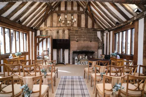 Coventry City Council A Tudor-looking room with brown beams and a slanted roof. It is laid out for a wedding ceremony with chairs on either side of an aisle. A rustic chandelier is hung from the ceiling.