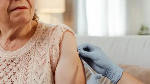 An elderly woman, her face only partially visible, is being given an injection by someone with blue, latex gloves on. The woman is wearing a short-sleeved woolen top.