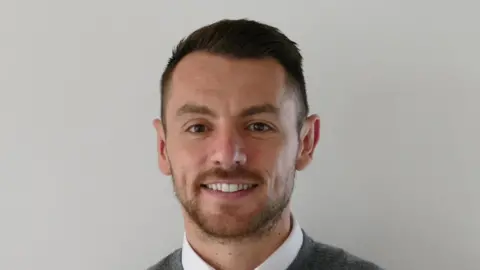 A man with dark hair smiling. He is wearing a grey jumpter and a white shirt.