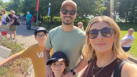 Emma Giannuzzi A man and woman wearing sunglasses are standing next to two boys and they are all smiling. Several people are walking on a footpath behind them.