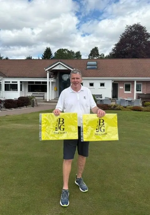 DOUGLAS SPENCER Douglas, stood on the grass outside the entrance to the Boat of Garten Golf Club. He is a man of average build with grey hair and is smiling. He's holding up two yellow flags and smiling.