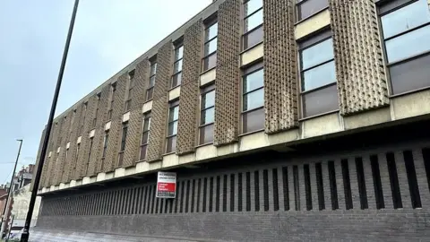A three-storey building with patterned concrete slabs along its front - each placed either side of a window. There is a red and white marketing sign fixed to the side of the building.