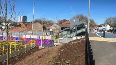 The new Willenhall station features colourful artwork and accessible lifts
A newly built railway station is seen from street level with a grey lift tower bearing the National Rail logo. Colourful purple and orange murals depicting cogs and flowers decorate the platform-level walls, and yellow accessibility ramps lead down to the platforms.